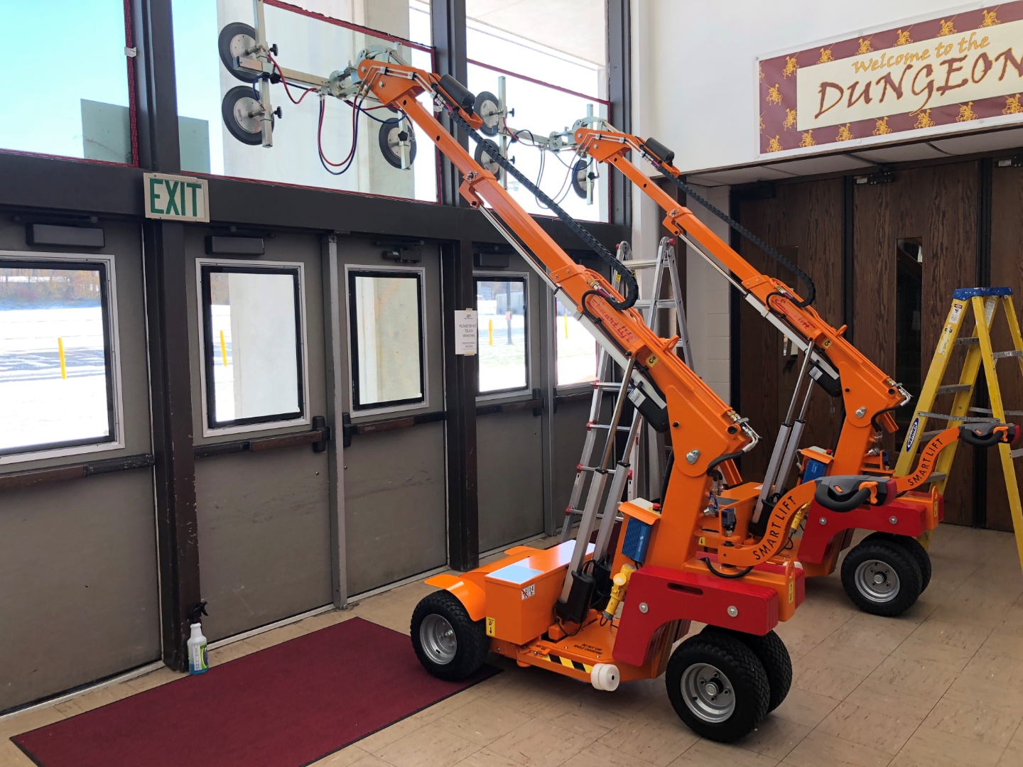 Two Smartlift glass lifters staged in a high school entry vestibule during a BallistiMAX install in Indiana, with snow visible through the existing entry doors.