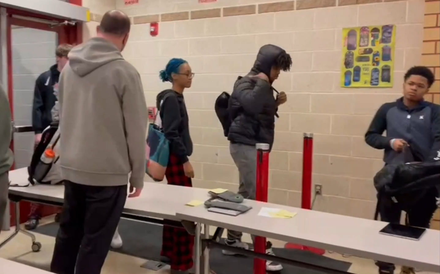 BallistiSCAN deployed at Penn Hills School District in Pittsburgh, Pennsylvania, 2023. Students walk single-file through a lane between two red stanchion-mounted sensor pucks at the front entrance, with a screening table holding confiscated phones and pocket items.