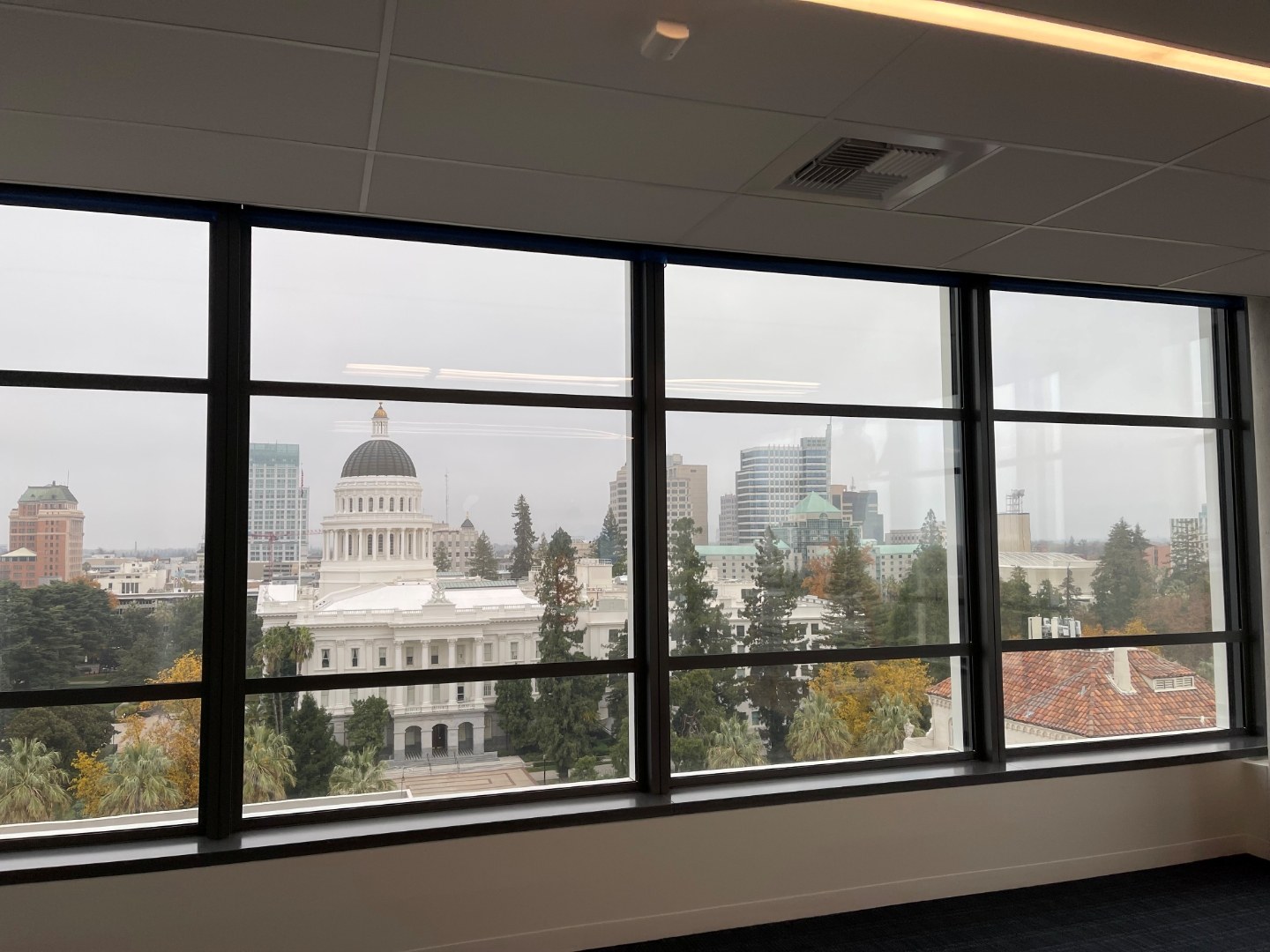 Floor-to-ceiling office windows on a high-floor government office in Sacramento, California, looking out across the State Capitol dome and the Sacramento skyline. The original glass remains in place; BallistiMAX panels are set back from the window line.