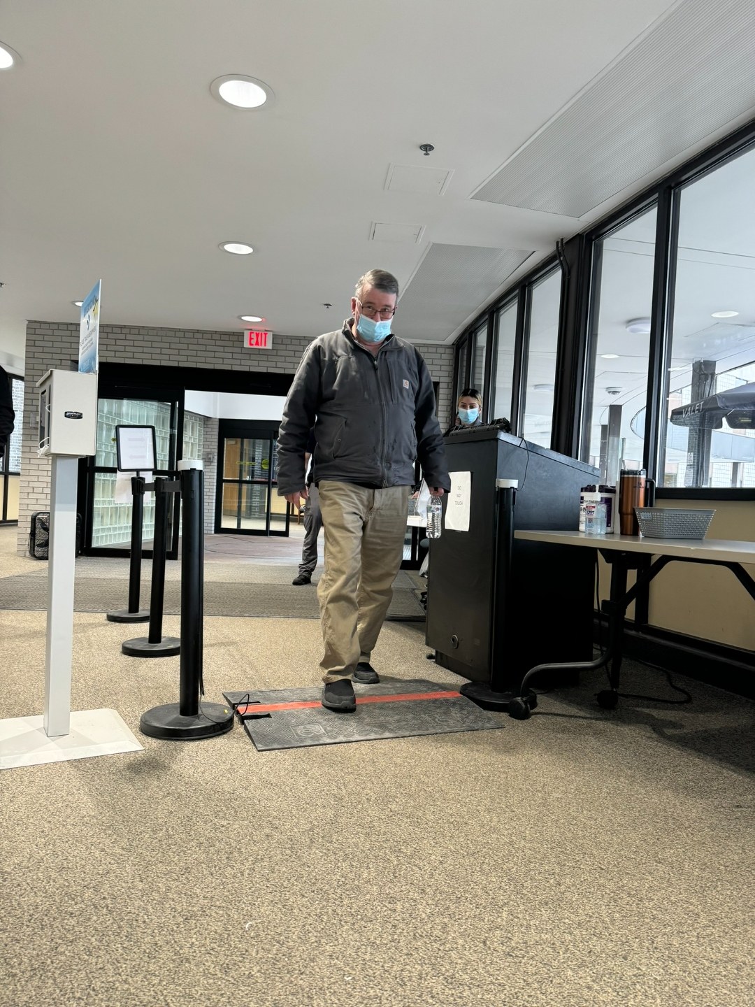 BallistiSCAN deployed at the public entrance of Upstate University Hospital in Syracuse, New York. A visitor in a face mask walks through the passive scan mat between two stanchion-mounted sensors, with the security desk and a state trooper visible to the side.