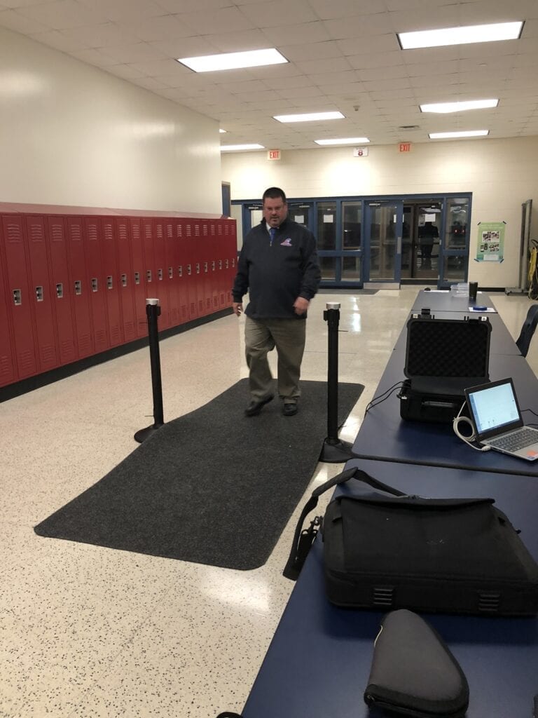 Man Walking Through A Metal Detector In A School Corridor